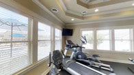 Located at 133 Boone Ridge Dr in Johnson City, Tennessee, this fitness center is a well-appointed, single-floor space designed to support a comprehensive workout routine. The room's most striking architectural feature is its elegant tray ceiling, accented with crisp white crown molding and outfitted with recessed lighting and a ceiling fan for comfortable air circulation during workouts. Multiple windows with slatted blinds line the walls, flooding the space with natural light and offering pleasant outdoor views of the surrounding neighborhood. A sliding glass door with blinds provides an additional connection to the outdoors. The flooring is covered with durable rubber gym matting, practical for both equipment placement and floor exercises. The space is equipped with a range of cardio machines, including a treadmill, elliptical trainer, stationary bike, and a rowing machine, alongside a weight bench and stability balls for strength and flexibility training. A wall-mounted flat-screen television is positioned for easy viewing from the cardio equipment, enhancing the workout experience. The warm, neutral-toned walls and thoughtful architectural details give this fitness space a refined, welcoming ambiance that goes well beyond a standard gym setting.