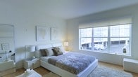 This single-floor space showcases a thoughtfully designed bedroom that emphasizes clean lines, neutral tones, and a calm, contemporary atmosphere. The room is anchored by a low-profile white platform bed dressed in layered neutral bedding — including textured throws and mixed decorative pillows — that contributes to the serene aesthetic. Matching white nightstands flank the bed, each topped with soft ambient lighting that adds warmth to the otherwise cool palette.

The walls are finished in crisp white, providing a bright, airy backdrop that is complemented by two framed coral-motif art pieces hung symmetrically above the headboard, introducing subtle organic detail. Natural light flows generously through a large multi-pane window fitted with a roller shade, offering views of an adjacent urban streetscape. The window serves as a functional focal point, connecting the interior to the outside environment.

Warm-toned hardwood flooring grounds the space, adding texture and contrast to the white surroundings, while a plush area rug beside the bed enhances comfort underfoot. Overall, the space presents a well-balanced, modern retreat with a cohesive design language centered on simplicity and light.