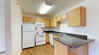 This functional kitchen space showcases a practical L-shaped design, maximizing efficiency in a compact area. Light-toned wood cabinetry lines the walls, providing ample storage with both upper and lower units. The cabinetry's natural finish adds warmth to the space, contrasting nicely with the white appliances.

The kitchen is equipped with essential white appliances, including a refrigerator, stove with oven, microwave, and dishwasher, all neatly integrated into the layout. Dark granite-like countertops offer durable work surfaces and add a touch of elegance to the overall design.

Underfoot, hardwood flooring in a medium tone complements the cabinetry and adds to the kitchen's inviting atmosphere. Overhead, a large fluorescent light fixture ensures bright, even illumination throughout the space.

The L-shaped configuration creates a small corner perfect for food preparation, while the adjacent counter space could serve as a casual dining area or additional workspace. Despite its compact size, this kitchen appears well-organized and designed for practical, everyday use.