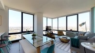 This thoughtfully designed space is situated on an upper floor, offering expansive panoramic city views through large, black-framed floor-to-ceiling windows that wrap around the corner of the unit. The windows flood the interior with abundant natural light, creating a bright and airy atmosphere throughout.

The open-plan layout seamlessly integrates the dining and living areas. The dining space features a sleek glass-top table paired with a wooden bench and teal accent chairs, adding a pop of color against the clean white walls and light hardwood flooring. A low-profile glass centerpiece with greenery adds a natural touch to the dining setting.

The adjoining living area is furnished with warm mustard-toned armchairs and a distinctive bronze metallic coffee table, which serves as a focal point on a striped area rug. A wall-mounted flat-screen television anchors the living space, positioned to take advantage of the city backdrop visible through the surrounding windows.

The overall ambiance is modern and sophisticated, characterized by a neutral palette accented with bold color choices, clean architectural lines, and an abundance of natural light enhanced by the striking elevated city views.