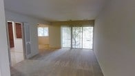This single-story space features a spacious, open living area finished with neutral wall-to-wall carpeting that creates a clean and understated aesthetic. The room is anchored by a large sliding glass door dressed with vertical blinds, which serves as the primary source of natural light and provides a direct view of mature trees and the outdoor surroundings beyond. The generous glazing allows daylight to filter throughout the interior, contributing to an airy and open atmosphere. A wide arched or squared doorway connects the main living area to an adjacent room, also carpeted and brightened by its own window, suggesting a flexible layout suitable for a variety of uses such as a bedroom, den, or dining area. The walls are finished in a light neutral tone, complementing the pale carpeting and maintaining a consistent, cohesive look throughout. Ceiling-mounted HVAC vents are visible, indicating a central air system. The overall design is straightforward and functional, with an emphasis on open sightlines, natural light, and a neutral color palette that provides a versatile foundation for furnishing and personalizing the space.