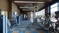 This well-appointed fitness center offers a thoughtfully designed workout environment spread across a single open floor. The space is anchored by a striking wall of floor-to-ceiling windows along one side, flooding the interior with an abundance of natural light and providing open outdoor views that create an energizing atmosphere during workouts. The flooring features a bold two-tone checkered pattern in blue and brown, lending the space a modern, polished aesthetic. Overhead, linear LED lighting fixtures and ceiling fans ensure consistent illumination and comfortable airflow throughout. On one side of the room, a row of cardio machines — including ellipticals and treadmills — lines the window wall, allowing users to exercise while enjoying the outdoor scenery. The opposite side is dedicated to strength training, with a well-organized arrangement of cable machines and multi-use weight equipment. A wall-mounted flat-screen television is positioned for easy viewing from the workout floor. Structural columns with metallic accents add an industrial design element that complements the space's contemporary finish. An entry door at the rear connects to an outdoor area, further enhancing the sense of openness and accessibility.