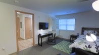 This single-floor space is designed with functionality and practicality in mind. The main room serves as both a living and sleeping area, featuring carpeted flooring accented by a green patterned area rug that adds a touch of color to the neutral palette of white walls and gray carpet. A wall-mounted flat-screen television anchors one side of the room, positioned above a dark-finished desk paired with a cushioned rocking chair, creating a dedicated workspace and entertainment zone. Adjacent to the desk, a white built-in shelving unit with open shelves and lower drawers provides ample storage and display space. A window fitted with horizontal blinds allows natural light to filter into the room, brightening the space. On the opposite side, a bed with neutral bedding is positioned alongside a dark bedside table. A doorway with natural wood trim leads to an adjacent area with light hardwood flooring, hinting at a connected hallway or additional room. The overall ambiance is clean, understated, and efficiently organized, making it well-suited for everyday living.