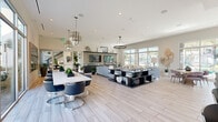 Located at 1902 Millenia Ave in Chula Vista, CA, this sophisticated single-story community clubhouse showcases a contemporary open-concept design finished with light wood-look tile flooring throughout. The main gathering area is anchored by elegant chandelier lighting — including a striking multi-tiered metallic fixture — and flows seamlessly between distinct functional zones. A shiplap-accented feature wall houses mounted flat-screen televisions above a sleek, dark-toned credenza, creating a polished media focal point. Adjacent to this is a well-appointed lounge area with plush gray seating and abundant greenery, bathed in natural light from large picture windows and glass sliding doors that open directly to the outdoor pool and patio. A dedicated dining area with a long white table and modern swivel chairs adds a collaborative gathering space. The fully equipped demonstration kitchen impresses with dark cabinetry, stainless steel appliances including a professional-grade range with hood and a full-size side-by-side refrigerator, and a dramatic marble slab backsplash complemented by neutral countertops. Vibrant abstract artwork and carefully curated décor complete the upscale aesthetic, while the seamless indoor-outdoor connection offers bright, resort-style living views throughout.