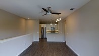This single-floor space showcases a thoughtfully designed open-concept layout that connects the living area and kitchen in a seamless, functional flow. Dark hardwood flooring runs throughout, adding warmth and contrast to the neutral, light-toned walls and crisp white cabinetry. The living area is anchored by a sleek ceiling fan with an integrated light fixture, while recessed lighting provides even, ambient illumination across the space. The kitchen is a standout feature, equipped with stainless steel appliances — including a refrigerator and microwave — set against a textured stone tile backsplash that adds visual depth and character. A peninsula counter with pendant lighting serves as a natural divide between the kitchen and living area, offering both a casual dining option and additional prep space. The pendant and track lighting above the kitchen add a modern, layered lighting aesthetic. Neutral wall tones throughout create a versatile backdrop suited to a variety of interior styles. The open sightlines between living and kitchen areas make the space feel expansive and well-connected, ideal for everyday living and entertaining alike.