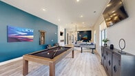 Located in Knoxville, TN, this single-floor community space offers a thoughtfully designed open-concept layout that seamlessly blends a kitchen, lounge, and game area into one cohesive environment. The entrance leads into a bright, welcoming interior finished with light wood-look flooring that flows throughout the entire space.

The kitchen and living area serve as the heart of the room, highlighted by a striking veined white quartz island with bar seating, grey cabinetry, and a dramatic floor-to-ceiling textured stone accent wall framing a mounted TV. Pendant lighting in mixed metallic finishes adds a modern, curated touch overhead, while a bold teal accent wall provides vibrant contrast.

The lounge zone features a built-in white paneled surround housing both a TV and a see-through fireplace, flanked by contemporary leather seating against a deep teal wall — creating an intimate, stylish retreat within the larger space. Adjacent, the game area is generously sized to accommodate a full pool table and foosball table, anchored by a colorful area rug and a decorative sputnik chandelier. Large windows along the perimeter flood the space with natural light and offer views of the surrounding landscaped community, enhancing the open and airy atmosphere throughout.