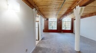 Located at 155 W Main St in Vernon Rockville, Connecticut, this two-floor loft-style space blends industrial heritage with residential comfort. The defining architectural character comes from the warm wood plank ceilings supported by heavy exposed timber beams, which run throughout both levels and create a rich, textured overhead canopy. Original exposed brick walls add to the historic charm, while large multi-pane windows flood the main living areas with abundant natural light and offer views of surrounding greenery.

The main living area is open and generously proportioned, with soft carpeting underfoot and white walls that contrast beautifully against the warm tones of the brick and wood. A staircase with a wooden handrail connects the two floors, with the upper level providing additional carpeted rooms, including one featuring an arched brick window and another with a built-in closet. A chandelier accents one of the living spaces, adding a touch of elegance.

The kitchen is equipped with oak-trimmed cabinetry, white countertops, a dishwasher, and a stainless steel sink, complemented by tile flooring. The bathroom includes a full tub-shower combination and tile flooring. Exposed piping and ductwork contribute to the loft's authentic industrial aesthetic throughout.