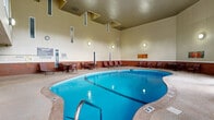 Located in Little Canada, MN, this generously sized indoor recreational facility offers an impressive array of amenities centered around a large, freeform swimming pool with striking blue water. The space spans a single floor and is characterized by soaring ceilings that create an open, airy atmosphere throughout. Tall, angled windows along one wall flood the pool area with abundant natural light while providing views of the surrounding landscape, and multiple glass-panel doors offer direct access to the outdoors.

The pool area is surrounded by a spacious aggregate concrete deck furnished with lounge chairs, seating tables, and dining-style chairs, providing ample room for relaxation. A lower brick wainscoting detail runs along the perimeter walls, accented by decorative artwork that adds a welcoming touch to the space.

Positioned along the poolside wall is an enclosed fitness center, visible through large picture windows. The gym is well-equipped with treadmills, ellipticals, and strength training equipment, offering convenient access directly from the pool deck. An upper-level mezzanine overlooks the pool area, adding architectural depth and vertical interest to the overall design. This facility is well-suited for year-round recreational use, combining aquatic amenities with fitness resources in one cohesive, well-appointed space.