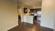 This single-floor residence showcases a contemporary open-concept layout centered around a well-appointed kitchen. Dark espresso cabinetry contrasts sharply with light countertops and a white peninsula that serves as both a workspace and a natural divider between the kitchen and living areas. The kitchen is equipped with a full suite of stainless steel appliances, including a refrigerator, electric range, and built-in microwave, making it both functional and visually cohesive.

Overhead, a combination of track lighting and pendant lights with warm-toned shades provides layered illumination throughout the space. The rich, dark hardwood flooring extends consistently across the main living areas, adding warmth and a sense of continuity to the open floor plan.

A hallway visible from the main living area leads to additional rooms, including a bathroom finished in complementary neutral tones. The overall design aesthetic is modern and streamlined, with clean lines, neutral wall colors, and carefully selected fixtures that contribute to a polished, cohesive interior. The space balances practicality with contemporary style throughout.