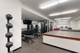 Located at 110 W Ocean Blvd in Long Beach, California, this fitness center is a fully equipped exercise room designed to support a comprehensive workout routine. The space is organized into distinct functional zones, making efficient use of its compact footprint.

The strength training area is anchored by a rack of rubber hex dumbbells, multiple cable and multi-use weight machines, and adjustable benches, all positioned alongside full-length mirrored walls that visually expand the room and allow users to monitor their form. A wall-mounted flat-screen television is positioned above the mirror for entertainment during workouts.

The cardio zone features treadmills and an elliptical trainer, each equipped with individual display consoles. A medicine ball storage rack with varying ball weights is also conveniently positioned nearby.

A raised, padded platform occupies a central portion of the room, bordered by a wood-trimmed edge, providing a dedicated area for stretching, yoga, or mat-based exercises. Steps provide easy access to the platform. Adjacent to this area is a dark storage cabinet stocked with fresh towels, adding a practical amenity for users.

Rubber flooring throughout ensures safety and durability, while bright overhead fluorescent lighting provides consistent, even illumination across the entire space. The clean white walls and organized layout contribute to a functional and professional fitness environment.