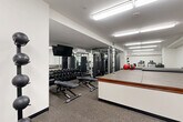 Located at 110 W Ocean Blvd in Long Beach, California, this fitness center is a fully equipped exercise room designed to support a comprehensive workout routine. The space is organized into distinct functional zones, making efficient use of its compact footprint.

The strength training area is anchored by a rack of rubber hex dumbbells, multiple cable and multi-use weight machines, and adjustable benches, all positioned alongside full-length mirrored walls that visually expand the room and allow users to monitor their form. A wall-mounted flat-screen television is positioned above the mirror for entertainment during workouts.

The cardio zone features treadmills and an elliptical trainer, each equipped with individual display consoles. A medicine ball storage rack with varying ball weights is also conveniently positioned nearby.

A raised, padded platform occupies a central portion of the room, bordered by a wood-trimmed edge, providing a dedicated area for stretching, yoga, or mat-based exercises. Steps provide easy access to the platform. Adjacent to this area is a dark storage cabinet stocked with fresh towels, adding a practical amenity for users.

Rubber flooring throughout ensures safety and durability, while bright overhead fluorescent lighting provides consistent, even illumination across the entire space. The clean white walls and organized layout contribute to a functional and professional fitness environment.