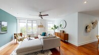 This thoughtfully designed single-floor residence blends modern aesthetics with warm, inviting materials throughout. Rich hardwood flooring flows seamlessly across every room, anchoring the space with a sense of warmth and continuity.

The living area is bright and airy, with multiple large windows framing expansive views of the surrounding cityscape and treetops. A ceiling fan overhead and a sectional sofa arranged around a round coffee table create a comfortable, relaxed atmosphere. A mid-century style credenza and circular wall mirror add refined character to the room.

Adjacent to the living space, the kitchen features sleek, high-gloss white cabinetry, a clean tile backsplash, and a full suite of stainless steel appliances, including a range, microwave, and dishwasher. The minimalist design keeps the space functional and uncluttered.

The dining area accommodates a full-sized table and chairs, with distinctive wall art adding visual interest. The space connects naturally to the rest of the home, supporting an open flow ideal for entertaining.

Two well-proportioned bedrooms each offer natural light through large windows with city and park views, ceiling fans, and warm hardwood floors. A dedicated home office provides a quiet retreat with its own window view.

The bathroom is finished with large-format grey tile, a floating wood-toned vanity, and a glass-enclosed walk-in shower, delivering a clean and contemporary feel throughout.