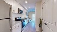 This thoughtfully designed single-floor studio presents a clean, modern aesthetic throughout its open layout. The galley-style kitchen runs along one wall and is outfitted with stainless steel appliances, including an electric range, over-the-range microwave, and dishwasher, complemented by crisp white cabinetry, granite-style countertops, and a double-basin sink. Light wood-style plank flooring flows seamlessly from the kitchen through the rest of the unit, creating a cohesive and airy feel. The living area is bathed in natural light through a floor-to-ceiling vertical blind-covered sliding glass door, which also provides access to the outdoors. The space is styled with a light blue sofa, a leaning shelf unit, and an area rug, contributing to the calm, neutral palette of the interior. The sleeping area is open to the main living space and accommodates a full-sized bed, a wardrobe cabinet, and a small desk arrangement, with a wall-mounted television adding convenience. Recessed lighting and a well-maintained bathroom with standard fixtures round out this efficiently appointed unit. The overall ambiance is bright, clean, and contemporary, making effective use of every square foot.