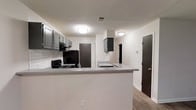 This single-floor, 3-bedroom, 2-bathroom home offers a well-organized layout designed to maximize comfort and functionality. The kitchen serves as a central feature of the space, showcasing gray cabinetry, a classic white subway tile backsplash, and a peninsula countertop that creates a natural divide between the kitchen and the adjacent living area. Black appliances, including a refrigerator and a gas range, lend a bold contrast to the neutral palette of white walls and gray countertops. Flush-mount ceiling lights provide consistent illumination throughout. The flooring transitions smoothly across the open areas with wood-look planks that add warmth and a modern touch to the interior. Multiple doorways visible from the kitchen connect seamlessly to the rest of the home, including the bedrooms and bathrooms, ensuring a logical and easy-to-navigate floor plan. The overall aesthetic is clean and contemporary, with a neutral color scheme that provides a versatile backdrop for a variety of interior styles. The combination of practical storage, open sightlines, and cohesive design elements makes this home a functional and inviting space suited for a range of lifestyles.