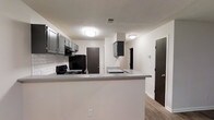 This single-floor, 3-bedroom, 2-bathroom home offers a well-organized layout designed to maximize comfort and functionality. The kitchen serves as a central feature of the space, showcasing gray cabinetry, a classic white subway tile backsplash, and a peninsula countertop that creates a natural divide between the kitchen and the adjacent living area. Black appliances, including a refrigerator and a gas range, lend a bold contrast to the neutral palette of white walls and gray countertops. Flush-mount ceiling lights provide consistent illumination throughout. The flooring transitions smoothly across the open areas with wood-look planks that add warmth and a modern touch to the interior. Multiple doorways visible from the kitchen connect seamlessly to the rest of the home, including the bedrooms and bathrooms, ensuring a logical and easy-to-navigate floor plan. The overall aesthetic is clean and contemporary, with a neutral color scheme that provides a versatile backdrop for a variety of interior styles. The combination of practical storage, open sightlines, and cohesive design elements makes this home a functional and inviting space suited for a range of lifestyles.