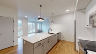 Located at 7900 SE Luther Rd in Portland, Oregon, this modern single-floor unit offers a thoughtfully designed open-concept layout with contemporary finishes throughout. The kitchen serves as the heart of the space, featuring a large center island with a white quartz countertop, gray shaker-style cabinetry, a black matte sink fixture, and stainless steel appliances including a refrigerator, electric range with over-the-range microwave, and dishwasher. Stylish pendant lights hang above the island, complementing the recessed lighting overhead. Light-toned hardwood flooring flows seamlessly throughout the entire unit, lending warmth and cohesion to every room. The spacious living area is bathed in natural light through large windows and offers direct access to an outdoor balcony, with views of the surrounding Portland neighborhood. The bedrooms are generously sized, each featuring large windows that frame tranquil outdoor scenery. The primary bathroom is appointed with a dual-sink vanity, navy blue cabinetry, white quartz countertops, and elegantly lit wall sconces above a wide mirror. A second full bathroom includes a tub-shower combination, dark vanity, and white countertop. Clean white walls and an abundance of natural light give the entire space a bright, airy, and move-in-ready feel.