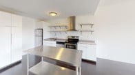 This well-appointed one-bedroom, one-bathroom unit at Dwell offers a thoughtfully designed single-floor layout that balances modern aesthetics with everyday functionality. The kitchen is a standout feature, equipped with stainless steel appliances including a refrigerator, electric range with a bold stainless steel range hood, and a dishwasher. White cabinetry and open stainless steel shelving provide ample storage, while the stainless steel island and countertops lend an industrial-modern edge. Rich, dark hardwood flooring flows through the main living areas, complemented by bold gray accent walls that create a contemporary, grounded atmosphere. Natural light filters in through the windows, enhancing the airy feel of the space. The bedroom features plush carpeting, crisp white walls, and a window fitted with blinds that allow for adjustable light and privacy. A closet area provides convenient built-in storage. The bathroom is clean and minimalist, showcasing large-format gray tile flooring, a dark wood vanity with a white countertop, and a full-size toilet, all framed by bright white walls. Throughout the unit, the design emphasizes clean lines, neutral tones, and practical finishes that come together to create a modern and comfortable living environment.