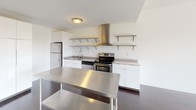 This well-appointed one-bedroom, one-bathroom unit at Dwell offers a thoughtfully designed single-floor layout that balances modern aesthetics with everyday functionality. The kitchen is a standout feature, equipped with stainless steel appliances including a refrigerator, electric range with a bold stainless steel range hood, and a dishwasher. White cabinetry and open stainless steel shelving provide ample storage, while the stainless steel island and countertops lend an industrial-modern edge. Rich, dark hardwood flooring flows through the main living areas, complemented by bold gray accent walls that create a contemporary, grounded atmosphere. Natural light filters in through the windows, enhancing the airy feel of the space. The bedroom features plush carpeting, crisp white walls, and a window fitted with blinds that allow for adjustable light and privacy. A closet area provides convenient built-in storage. The bathroom is clean and minimalist, showcasing large-format gray tile flooring, a dark wood vanity with a white countertop, and a full-size toilet, all framed by bright white walls. Throughout the unit, the design emphasizes clean lines, neutral tones, and practical finishes that come together to create a modern and comfortable living environment.