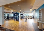 Located in Flagler Village at 405 NE 2nd Street in Fort Lauderdale, Florida, this comprehensive fitness center is designed to support a full range of workout routines across a generously sized, single-floor layout. The space features warm hardwood-style flooring throughout the main workout areas, complemented by rubber flooring in the strength and stretching zones, creating both a polished and functional environment.

The cardio zone is lined with floor-to-ceiling windows that invite abundant natural light and offer views of the surrounding palm-lined landscape, housing a row of modern treadmills, ellipticals, and rowing machines. Adjacent to this area, a well-stocked free weights section features a full dumbbell rack, barbell stations with squat racks, and a large mirrored wall, catering to a wide range of strength training needs.

The functional training area includes a cable machine system, a punching bag, cycling bikes, and additional specialty equipment. Pendant lighting throughout adds an elevated aesthetic to the industrial-modern design scheme accented by deep teal and charcoal wall tones.

A separate, dedicated studio room provides space for yoga, Pilates, and group fitness activities, complete with exercise mats, medicine balls, resistance equipment, and full-length mirrors. A hydration station with water fountains and bottled water storage ensures convenience throughout the workout experience.