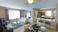 This single-floor residence offers an open-concept layout that seamlessly connects the living, dining, and kitchen areas into one cohesive and functional space. The living room is generously sized, anchored by a plush white sofa, a glass-top coffee table, and a soft area rug, all bathed in natural light streaming through large windows dressed with neutral curtains. The airy, neutral color palette throughout lends the space a calm and welcoming ambiance. Adjacent to the living area, a dedicated dining space accommodates a dark-toned dining table with cushioned chairs, ideal for everyday meals or entertaining guests. The kitchen is outfitted with stainless steel appliances, including a full-size refrigerator, rich dark wood cabinetry, and a white countertop breakfast bar complete with bar seating. A pendant light adds a refined touch above the bar area. A sliding glass door beyond the living room hints at access to an outdoor area, further enhancing the sense of openness. The smooth, light-toned walls and consistent flooring throughout create a unified, well-designed interior that balances comfort with practicality.
