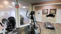 This single-level fitness center offers a practical and functional workout space equipped with essential cardio machines, including a prominent elliptical trainer and a treadmill positioned along the perimeter. The room features patterned carpet flooring and warm, neutral-toned walls that create an understated and focused exercise environment. A standout architectural detail is the glass-paneled partition wall, which separates the main equipment area from an adjoining enclosed exercise room. This secondary space, visible through the frosted glass panels, appears to feature hard flooring suitable for stretching, yoga, or floor-based workouts. A clearly posted "Fitness" sign with rules and guidelines is displayed at the entrance to this glass room, maintaining an organized and informative atmosphere. An additional door on the far wall likely leads to a restroom or utility area, adding convenience to the facility. Overhead lighting provides adequate illumination throughout. The overall layout is efficient and straightforward, designed to accommodate a range of fitness activities within a compact, well-organized space.