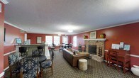 This single-floor space functions as a welcoming communal gathering area, thoughtfully designed to encourage both relaxed socializing and casual dining. The room is anchored by a prominent brick fireplace with a white mantle, flanked by decorative ceramic urns and framed artwork, lending a classic, homey character to the space. Warm terracotta-red walls contrast with white crown molding and window trim, creating a visually rich yet cohesive interior palette. The textured ceiling adds architectural dimension throughout.

Natural light filters in through multiple double-hung windows along the far wall, brightening a dining area furnished with traditional wooden tables and chairs. A bar-height counter with tiled detailing and bar stools defines a separate casual seating zone, ideal for light refreshments or conversation. The central lounge area features a leather sofa and an upholstered ottoman arranged around a patterned area rug, creating an inviting focal point. Recessed and pendant lighting provide consistent illumination throughout, ensuring the space remains functional and comfortable during evening hours.
