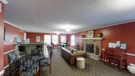 This single-floor space functions as a welcoming communal gathering area, thoughtfully designed to encourage both relaxed socializing and casual dining. The room is anchored by a prominent brick fireplace with a white mantle, flanked by decorative ceramic urns and framed artwork, lending a classic, homey character to the space. Warm terracotta-red walls contrast with white crown molding and window trim, creating a visually rich yet cohesive interior palette. The textured ceiling adds architectural dimension throughout.

Natural light filters in through multiple double-hung windows along the far wall, brightening a dining area furnished with traditional wooden tables and chairs. A bar-height counter with tiled detailing and bar stools defines a separate casual seating zone, ideal for light refreshments or conversation. The central lounge area features a leather sofa and an upholstered ottoman arranged around a patterned area rug, creating an inviting focal point. Recessed and pendant lighting provide consistent illumination throughout, ensuring the space remains functional and comfortable during evening hours.