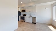 This single-floor unit features a clean, contemporary design characterized by white walls, light wood-style flooring, and an open layout that connects the main living areas seamlessly. The living space is generously proportioned and bathed in natural light through large windows that frame expansive city and treetop views, complemented by a ceiling fan overhead. The open-concept kitchen is efficiently designed with white flat-panel cabinetry extending to the ceiling, stainless steel appliances including a range, microwave, dishwasher, and refrigerator, along with a single-basin sink and ample counter space. Recessed lighting illuminates the kitchen area, maintaining a clean and modern aesthetic. The bedroom offers a comfortable footprint with warm wood-style flooring and access to a well-designed walk-in closet featuring built-in shelving, hanging rods, and dedicated storage compartments, providing excellent organizational capacity. The bathroom is finished with neutral vinyl flooring, a full tub-shower combo with a white surround, and clean, simple fixtures throughout. The unit's overall palette of whites and warm neutrals creates a bright, cohesive ambiance throughout.