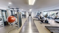 The Wellington Fitness Center in Arlington, Virginia is a spacious, single-floor facility designed to accommodate a full range of workout needs. The fitness center is organized into distinct zones, creating an efficient and functional layout throughout the open floor plan.

The cardio area is well-stocked with Matrix-brand treadmills, elliptical trainers, stair climbers, stationary bikes, and rowing machines, arranged in orderly rows to maximize space and accessibility. Large windows along one wall allow natural light to fill portions of the space, offering views to the outdoors and contributing to an open, energizing atmosphere.

The strength training section features a comprehensive selection of selectorized cable machines, multi-functional weight stations, adjustable benches, and a tiered dumbbell rack with a full range of free weights. Mirrored walls throughout the facility enhance the sense of space and allow users to monitor their form during exercise.

The flooring transitions between durable hardwood-style planks in central walkway areas and rubber-composite surfaces in equipment zones, providing both visual contrast and practical functionality. Recessed and panel lighting ensures consistent illumination throughout the entire space. The overall ambiance is clean, modern, and purposefully designed to support a productive fitness experience.