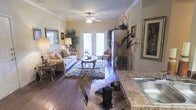 This single-floor space offers a warm and inviting atmosphere, highlighted by rich hardwood flooring that flows throughout the main living area. The living room is tastefully appointed, featuring crown molding, a ceiling fan with a light fixture, and soft neutral-toned walls that create a cozy ambiance. A plush area rug anchors the seating arrangement, which centers around an elegant oval coffee table. Lush indoor plants and framed wall art add character and a welcoming touch to the space. Natural light pours in through a set of classic French doors at the far end of the room, which also provide a view to the outside and lend an airy quality to the interior. A cushioned armchair positioned near the doors offers a peaceful reading nook. Adjacent to the living area, the kitchen showcases granite countertops with warm, varied toning, along with a stainless steel sink. The open sightlines between the kitchen and living room enhance the sense of spaciousness, making this a functional and aesthetically cohesive single-level residence.