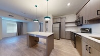 This single-floor unit showcases a contemporary design with light hardwood-style flooring that runs consistently throughout the space, creating a cohesive and clean aesthetic. The open-concept kitchen serves as a central feature, equipped with stainless steel appliances including a refrigerator, electric range, microwave, and dishwasher. Warm-toned, textured cabinetry with matte black hardware provides ample storage, complemented by white countertops and a stainless steel sink. A central kitchen island with a white surface and built-in outlets doubles as a workspace or dining area, illuminated by two teal pendant lights that add a distinctive pop of color.

Adjacent to the kitchen, an in-unit stacked washer and dryer adds everyday convenience. The living area is bright and open, featuring neutral walls and a window dressed with taupe drape panels. The bedroom continues the light, airy feel with a similarly styled window allowing natural light to filter in. Storage is well-addressed through a dedicated closet area featuring both hanging rods and multiple open shelving tiers. The bathroom is finished with a wood-toned vanity, white quartz countertop, open shelving, and a backlit mirror, reflecting the unit's consistent modern sensibility.