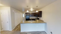 This single-floor residence offers a thoughtfully designed open-concept layout that seamlessly connects the kitchen and living areas. The kitchen is a standout feature, showcasing rich dark cabinetry that contrasts elegantly with light-toned countertops. A peninsula counter serves as both a functional workspace and a natural divider between the kitchen and living space, making it ideal for casual dining or entertaining. Two stylish pendant lights hang above the peninsula, adding warm, focused illumination to the area. The kitchen is fully equipped with black appliances, including a refrigerator, range, and microwave, along with a double-basin sink set into the peninsula. Ample upper and lower cabinet storage ensures practical organization throughout. The flooring transitions from wood-look planks in the kitchen and entry areas to carpet in the living space, adding warmth and texture underfoot. Neutral wall tones and white trim throughout the space create a clean, bright ambiance. A closet with bifold doors is conveniently located near the entryway, and a door with a sidelight window provides natural light at the rear of the space. The overall design balances functionality with a modern, cohesive aesthetic well-suited for everyday living.