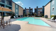 This outdoor amenity space centers around a generously sized swimming pool with clear, blue-green water, offering a refreshing retreat for residents. The pool is surrounded by a wide concrete deck that provides ample room for relaxation and movement. Multiple lounge chairs are arranged along the poolside, creating a comfortable setting for sunbathing or leisure.

The pool area is framed by multi-story residential buildings finished in teal and dark navy tones, giving the complex a cohesive and modern aesthetic. Brick-accented detailing and vertical architectural columns add structure and visual interest to the building facades. Balconies on the upper floors overlook the pool courtyard, contributing to a sense of community within the space.

Mature trees and neatly trimmed shrubs are interspersed throughout the courtyard, softening the hardscape and providing pockets of shade and greenery. The surrounding landscaping enhances the overall ambiance, making the outdoor space feel inviting and well-kept. The area benefits from abundant natural light, with open sky views that contribute to a bright, airy atmosphere throughout the day.
