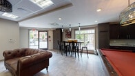 Located at 10 Kings Rd, this single-floor amenity lounge is thoughtfully designed to offer a variety of social and recreational spaces within one cohesive environment. The layout flows naturally between distinct zones, each with its own character while maintaining a unified contemporary aesthetic throughout.

The main open area is anchored by a striking red-felt pool table set against a textured geometric feature wall, illuminated by a bold metallic pendant light overhead. Dark cabinetry with a wet bar lines one wall, adding functional convenience to the games area. Large windows along the perimeter flood the space with natural light and provide views of the surrounding streetscape and greenery outside.

A bar-height dining table surrounded by black industrial-style stools sits adjacent to the windows, topped by slender cylinder pendant lights that add a modern touch. The area connects seamlessly to the main entrance, which features a glass door offering additional views and natural light.

A separate, more intimate lounge area provides a quieter retreat, furnished with a deep brown tufted leather Chesterfield sofa and cream wingback chairs arranged around a minimalist coffee table. A wall-mounted flat-screen television and an ornate ceiling fixture lend this nook a refined, comfortable ambiance. Light tile flooring and neutral wall finishes tie all areas of the space together cohesively.