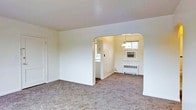 This single-floor space presents a clean, understated interior defined by its neutral white walls and wall-to-wall carpeting throughout. The main living area is open and uncluttered, offering a blank canvas with smooth ceilings and simple, functional design. A distinctive arched doorway serves as the central architectural feature, creating a graceful transition between the living area and an adjoining room. This arch adds a subtle classic character to an otherwise straightforward layout. The adjoining room receives natural light through a window, brightening the space and providing views of the outdoors. A ceiling-mounted light fixture in this secondary room offers additional illumination. Practical amenities are visible, including a wall-mounted air conditioning unit and a baseboard radiator, suggesting year-round climate control capability. A door visible along the wall of the main room leads to additional areas of the unit. The overall ambiance is quiet and neutral, making the space well-suited for a variety of uses. The consistent carpeting and coordinated white tones throughout both rooms lend a cohesive, move-in-ready feel to the interior.