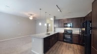 This single-floor residence showcases a thoughtfully designed open-concept layout that seamlessly connects the kitchen and living areas. The kitchen is a standout feature, equipped with rich dark-toned cabinetry that provides ample storage space, complemented by light granite countertops that add a touch of elegance. A central kitchen island with an integrated sink serves as both a functional workspace and a natural gathering point, illuminated by a trio of modern pendant lights that hang at varying heights, creating a visually appealing focal point. Black appliances, including a range and over-the-range microwave, lend a sleek, cohesive aesthetic to the culinary space. Track lighting along the kitchen ceiling ensures excellent task illumination, while a ceiling fan with a decorative fixture provides comfort and ambient light to the adjacent living area. The transition from hardwood-style flooring in the kitchen to plush carpeting in the living space adds warmth and definition between the two zones. Neutral wall tones throughout create a clean, versatile backdrop that suits a variety of interior styles. The open flow between spaces enhances the overall sense of roominess, making this residence both practical and inviting.
