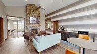 The Community Room at 3600 Meadow Dr in Grand Forks, North Dakota is a thoughtfully designed single-floor facility offering a generous and versatile layout. The living room serves as the heart of the space, featuring soaring ceilings, light wood-style flooring, and a striking floor-to-ceiling stacked stone fireplace with a wood mantel and wall-mounted TV above. Contemporary seating including a sage green sofa and tufted accent chairs surrounds a geometric wood coffee table, creating a warm and inviting atmosphere. A textured striped accent wall adds visual depth, while a large interior window provides a direct sightline into the fitness center beyond. The exercise room is spacious and well-lit, benefiting from multiple windows offering outdoor views of the surrounding greenery and abundant natural light supplemented by recessed ceiling panels. It is outfitted with a comprehensive selection of cardio equipment, including treadmills and ellipticals, as well as professional-grade Hoist strength machines. The kitchen and dining area features dark cabinetry with light countertops, a full suite of stainless steel appliances, a white subway tile backsplash, and a breakfast bar with seating. Light wood-style flooring and pendant lighting complete the modern, functional aesthetic. A connecting hallway ties the spaces together seamlessly.