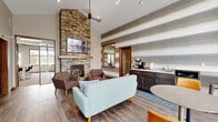 The Community Room at 3600 Meadow Dr in Grand Forks, North Dakota is a thoughtfully designed single-floor facility offering a generous and versatile layout. The living room serves as the heart of the space, featuring soaring ceilings, light wood-style flooring, and a striking floor-to-ceiling stacked stone fireplace with a wood mantel and wall-mounted TV above. Contemporary seating including a sage green sofa and tufted accent chairs surrounds a geometric wood coffee table, creating a warm and inviting atmosphere. A textured striped accent wall adds visual depth, while a large interior window provides a direct sightline into the fitness center beyond. The exercise room is spacious and well-lit, benefiting from multiple windows offering outdoor views of the surrounding greenery and abundant natural light supplemented by recessed ceiling panels. It is outfitted with a comprehensive selection of cardio equipment, including treadmills and ellipticals, as well as professional-grade Hoist strength machines. The kitchen and dining area features dark cabinetry with light countertops, a full suite of stainless steel appliances, a white subway tile backsplash, and a breakfast bar with seating. Light wood-style flooring and pendant lighting complete the modern, functional aesthetic. A connecting hallway ties the spaces together seamlessly.