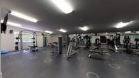 This fitness center is a comprehensively outfitted facility designed to accommodate a full range of workout routines. The space is organized across a single floor, with distinct zones dedicated to cardiovascular training and strength work. The cardio area features treadmills, elliptical machines, and a stair climber, all positioned in front of wall-mounted flat-screen televisions for an engaging workout experience. Strength training is well represented with a multi-station cable machine, a Smith machine, a dedicated bench press setup, and a tiered dumbbell rack stocked with a wide selection of free weights. Additional equipment includes various selectorized strength machines targeting specific muscle groups, a rowing machine, and balance tools such as a BOSU ball. The perimeter walls are lined with large mirrors that visually expand the space and allow users to monitor their form. The dark rubber flooring provides durability and safety underfoot, while recessed and overhead fluorescent lighting ensures the entire area is well illuminated. A teal accent wall near the entry adds a pop of color, and wall-mounted speakers contribute to the overall environment. A window in one corner allows for a touch of natural light, rounding out a practical and functional fitness facility.