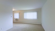 This single-floor space presents a clean, minimalist living area characterized by its neutral color palette and straightforward layout. The room features wall-to-wall carpeting in a light beige tone, contributing to a soft, understated aesthetic throughout. Two windows dressed with white vertical blinds line the exterior wall, allowing for flexible control of natural light while maintaining a bright and airy atmosphere. A window-unit air conditioner is installed beneath the smaller of the two windows, providing climate control alongside the baseboard heating system that runs along the base of the exterior wall — a practical combination for year-round comfort. The walls are finished in a light cream tone, complementing the overall neutral scheme of the space. Ceiling height appears standard, and a ceiling-mounted light fixture provides overhead illumination. The open layout of the room offers versatile arrangement possibilities, making it adaptable to various furniture configurations. The space maintains a simple, functional character with no ornamental architectural detailing, emphasizing practicality and ease of use.