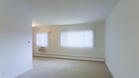 This single-floor space presents a clean, minimalist living area characterized by its neutral color palette and straightforward layout. The room features wall-to-wall carpeting in a light beige tone, contributing to a soft, understated aesthetic throughout. Two windows dressed with white vertical blinds line the exterior wall, allowing for flexible control of natural light while maintaining a bright and airy atmosphere. A window-unit air conditioner is installed beneath the smaller of the two windows, providing climate control alongside the baseboard heating system that runs along the base of the exterior wall — a practical combination for year-round comfort. The walls are finished in a light cream tone, complementing the overall neutral scheme of the space. Ceiling height appears standard, and a ceiling-mounted light fixture provides overhead illumination. The open layout of the room offers versatile arrangement possibilities, making it adaptable to various furniture configurations. The space maintains a simple, functional character with no ornamental architectural detailing, emphasizing practicality and ease of use.