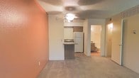 This single-floor residence offers a practical and comfortable layout designed for easy everyday living. The open living area is finished with neutral-toned carpet flooring and features a warm terracotta accent wall that adds a touch of color and personality to the space. A ceiling fan with a light fixture is centrally mounted, providing both air circulation and ambient lighting throughout the main area.

The kitchen is open to the living space and includes a peninsula-style breakfast bar with a laminate countertop, offering a casual dining or prep area. A white refrigerator and dark wood upper cabinetry are visible, along with a sink, making the kitchen functional and accessible. The open sightlines between the kitchen and living area create a connected, airy feel.

Just beyond the kitchen, a hallway leads to a full bathroom, which is visible from the main living space. The bathroom features light-toned finishes, a vanity with cabinetry, and a bathtub, maintaining the clean and neutral aesthetic found throughout the residence. The textured ceiling adds a classic finish consistent across all areas of the space, contributing to a cohesive interior design throughout.