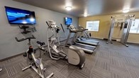 This fitness center offers a practical and comfortable environment for cardiovascular workouts. The space is outfitted with multiple treadmills, Precor elliptical trainers, and a stationary bike, providing users with a solid variety of cardio equipment to suit different exercise preferences. Wall-mounted flat-screen televisions are positioned throughout the room, allowing members to stay entertained during their workouts. The flooring consists of durable, patterned carpet tiles well-suited for a fitness environment. Overhead flush-mount lighting ensures the space is well-lit, while a window along one wall allows natural light to filter in, adding a sense of openness to the room. The walls are painted in a combination of cool grey and warm yellow tones, creating a clean and energizing atmosphere. A baseboard heater beneath the window suggests the space is designed for year-round comfort. Overall, this fitness center is a functional and focused facility that provides essential cardio equipment in a tidy, accessible setting.