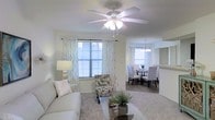 This single-story residence showcases an open-concept layout that seamlessly connects the living and dining areas, creating a spacious and airy feel. The living room is finished with plush carpeting in a warm neutral tone, providing a comfortable foundation for the space. A ceiling fan with an integrated light fixture serves as a functional centerpiece overhead, while sheer patterned curtains frame the windows, allowing generous natural light to filter through while maintaining a soft, inviting ambiance.

The living area flows naturally into the dining space, which is anchored by an elegant chandelier that adds a refined touch. The dining area features ample room for a full table and chairs, making it well-suited for both everyday meals and entertaining. Additional natural light enters through a second window in the dining area, further brightening the open space.

A pass-through architectural detail, visible between the living and dining areas, hints at a connected kitchen beyond, enhancing the home's cohesive, open feel. The overall palette of soft whites, creams, and grays creates a clean, contemporary aesthetic throughout, making this a welcoming and well-designed single-floor living space.