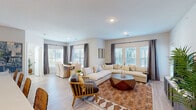 Located at 6407 E 163rd St in Belton, Missouri, this thoughtfully designed single-story home offers a cohesive, modern aesthetic with light-toned hardwood-style flooring and a neutral color palette flowing throughout. The open-concept living and dining area is bright and welcoming, with an abundance of windows dressed in elegant draping curtains that invite generous natural light. The living room comfortably accommodates a large sectional sofa and features a stylish geometric coffee table, creating an ideal space for both relaxing and entertaining. The adjacent dining area offers seating for multiple guests and is framed by large windows with outdoor views. The kitchen is a standout feature, centered around an expansive island with a white quartz countertop, undermount sink, and seating for several bar stools. Contemporary pendant lighting, warm wood-toned cabinetry, and stainless steel appliances complete the space. The primary bedroom is generously proportioned, carpeted for comfort, and includes a ceiling fan and ample natural light through multiple windows. Additional bedrooms are well-sized and versatile, with one configured as a dedicated home office featuring a closet and bright window. The bathroom showcases clean lines with wood-tone vanity cabinetry, white quartz counters, matte black fixtures, and a tiled tub-shower combination, reflecting the home's consistently modern design sensibility.