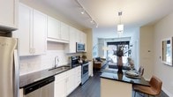This single-floor space showcases a thoughtfully designed open-concept layout that seamlessly connects the kitchen and living area. The kitchen is a standout feature, outfitted with crisp white shaker-style cabinetry, dark granite countertops, and a classic white subway tile backsplash that adds a clean, timeless aesthetic. Stainless steel appliances — including a range, microwave, and dishwasher — provide a sleek, modern touch, while a double-basin undermount sink with a contemporary faucet adds to the functionality. Track lighting runs along the kitchen ceiling, ensuring ample illumination throughout the workspace. A kitchen island with matching dark countertop provides additional prep space and casual seating. Rich dark hardwood-style flooring flows continuously from the kitchen into the living area, grounding the space with warmth and cohesion. The living area benefits from a large window that invites abundant natural light and offers views to the exterior, creating an airy, open ambiance. A pendant light fixture above the island bridges the two spaces stylistically, reinforcing the unit's modern, well-appointed character throughout.