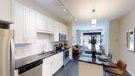 This single-floor space showcases a thoughtfully designed open-concept layout that seamlessly connects the kitchen and living area. The kitchen is a standout feature, outfitted with crisp white shaker-style cabinetry, dark granite countertops, and a classic white subway tile backsplash that adds a clean, timeless aesthetic. Stainless steel appliances — including a range, microwave, and dishwasher — provide a sleek, modern touch, while a double-basin undermount sink with a contemporary faucet adds to the functionality. Track lighting runs along the kitchen ceiling, ensuring ample illumination throughout the workspace. A kitchen island with matching dark countertop provides additional prep space and casual seating. Rich dark hardwood-style flooring flows continuously from the kitchen into the living area, grounding the space with warmth and cohesion. The living area benefits from a large window that invites abundant natural light and offers views to the exterior, creating an airy, open ambiance. A pendant light fixture above the island bridges the two spaces stylistically, reinforcing the unit's modern, well-appointed character throughout.