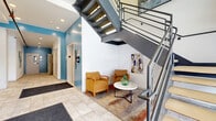Located at 2828 Euclid Avenue in Cleveland, Ohio, this two-floor amenity space offers residents a well-appointed selection of shared facilities. The ground floor serves as the primary entry point, featuring a bright, welcoming hallway finished with large-format tile flooring and accented by bold teal walls. A cozy seating nook with mustard-yellow chairs and a round side table sits beneath a striking open-tread metal staircase with industrial-style mesh railings, lending the space a modern, urban character. Artwork adorns the walls alongside practical building elements such as exposed fire suppression piping, adding to the building's distinctive aesthetic. An elevator provides convenient access between floors. The upper floor hosts the fitness center, a generously sized room outfitted with cardio equipment including treadmills, elliptical machines, and stationary bikes, as well as strength-training benches and a power rack. Wall-mounted televisions and large mirrors enhance the workout experience, while recessed lighting and carpeted flooring keep the space comfortable and functional. Exposed ductwork on the ceiling adds an industrial touch consistent with the building's overall design language. A dedicated laundry room rounds out the amenities, providing added convenience for building residents.