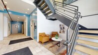 Located at 2828 Euclid Avenue in Cleveland, Ohio, this two-floor amenity space offers residents a well-appointed selection of shared facilities. The ground floor serves as the primary entry point, featuring a bright, welcoming hallway finished with large-format tile flooring and accented by bold teal walls. A cozy seating nook with mustard-yellow chairs and a round side table sits beneath a striking open-tread metal staircase with industrial-style mesh railings, lending the space a modern, urban character. Artwork adorns the walls alongside practical building elements such as exposed fire suppression piping, adding to the building's distinctive aesthetic. An elevator provides convenient access between floors. The upper floor hosts the fitness center, a generously sized room outfitted with cardio equipment including treadmills, elliptical machines, and stationary bikes, as well as strength-training benches and a power rack. Wall-mounted televisions and large mirrors enhance the workout experience, while recessed lighting and carpeted flooring keep the space comfortable and functional. Exposed ductwork on the ceiling adds an industrial touch consistent with the building's overall design language. A dedicated laundry room rounds out the amenities, providing added convenience for building residents.