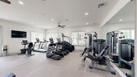 Located in Surf City, NC, this fitness center is thoughtfully designed to accommodate a wide range of workout needs across two functional zones. The main exercise area features neutral-toned carpet tiles and is lined with multiple windows that allow natural light to fill the space, complemented by ceiling fans and recessed lighting overhead. This zone is outfitted with a robust selection of TRUE-brand cardio equipment, including treadmills, ellipticals, and stair climbers, as well as a cable strength machine, a stationary bike, and a flat weight bench. A wall-mounted television and a large mirror add to the functionality of the room. Double glass-paned doors open to outdoor views, enhancing the bright and airy atmosphere. A separate studio room is distinguished by light hardwood flooring, floor-to-ceiling mirrored walls, and additional ceiling fans. This space is equipped for functional and group fitness, featuring a stationary bike, balance trainers, stability balls, and a wall-mounted display. The two areas are connected via a glass-paneled interior door, creating a seamless flow throughout the facility. The overall design is clean and modern, with white walls and an open layout that promotes an inviting, motivating environment.